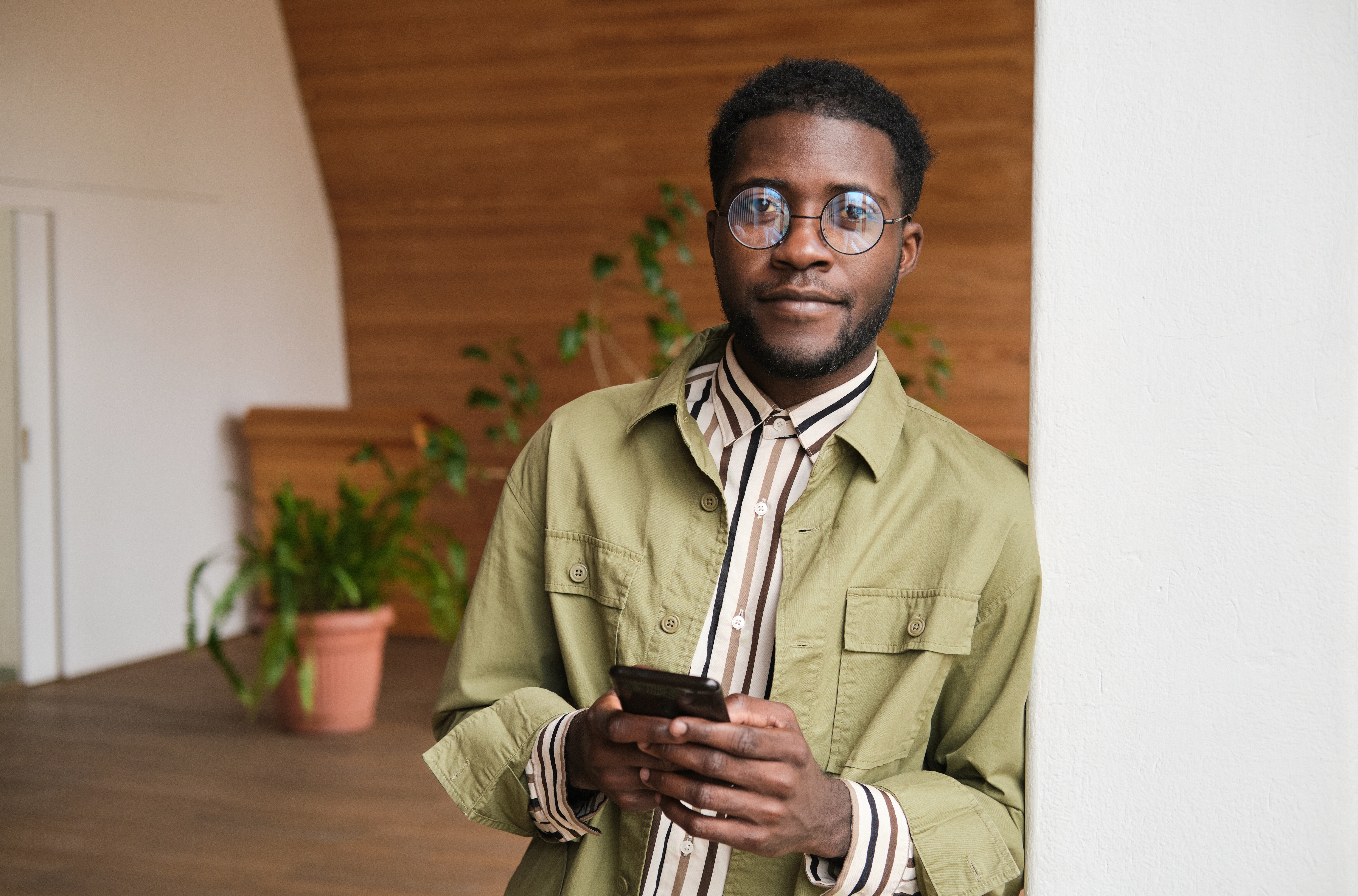 Portrait of African young businessman looking at camera and using mobile phone while standing at office